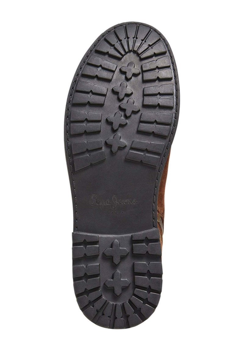 La suela del zapato presenta un material de goma con una superficie texturizada, surcos en un patrón y un diseño circular en el centro. Se aprecia la parte superior de color marrón.