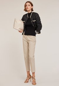 Schwarze gepolsterte Bomberjacke mit cremefarbenen Akzenten, schwarzer gerippter Pullover, beiger maßgeschneiderter Anzughose und sandfarbene, aufregende Absatzsandalen. Hält eine beige Tasche.