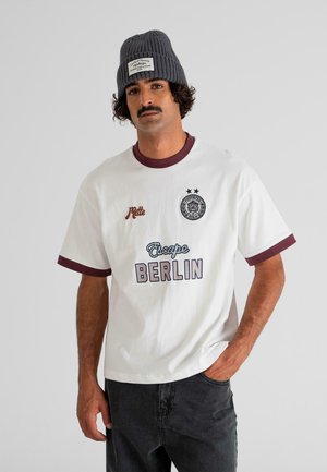 Mand med skæg iført en grå strikket hue og hvid T-shirt med teksten "Escape Berlin", stående med den ene hånd i lommen på sine sorte jeans.