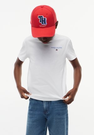 Persona con una gorra roja de Tommy Hilfiger y una camiseta blanca con un pequeño logo de Tommy Hilfiger, combinada con vaqueros azules, ajustando el dobladillo de la camiseta.