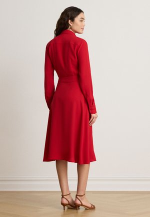 Femme aux longs cheveux foncés portant une robe midi rouge à manches longues et des sandales à talons beige, debout sur un sol en bois.