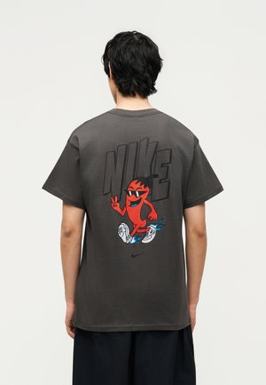 Άτομο που φοράει σκούρο γκρι μπλουζάκι Nike με μεγάλο κείμενο "NIKE" και ένα κόκκινο καρτούν χαρακτήρα με γυαλιά ηλίου και αθλητικά παπούτσια στην πλάτη.