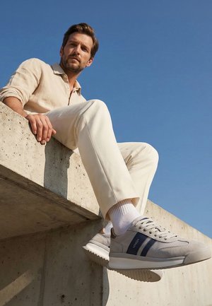 Mann in hellbeigem Outfit und weißen Sneakers mit blauen Streifen sitzt auf einer Betonmauer vor klarem blauem Himmel.