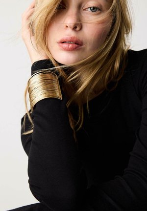 Donna bionda con maglia nera a maniche lunghe e grande bracciale dorato a polsino, appoggiando il viso sulla mano, ciocche di capelli sul volto.