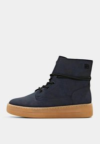 Stivaletti alla caviglia in suede blu navy con punta tonda, suola in gomma testurizzata e lacci neri. Presentano un logo sul lato esterno e un design snodato.