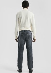 Mężczyzna w białej bluzie z długim rękawem, ciemnoszarych dżinsach i czarnych butach, stojący tyłem do kamery na jednolitym tle.