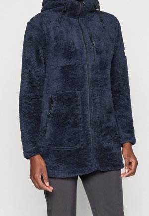 Persona que lleva una chaqueta azul marino, difusa, con cremallera, capucha y bolsillos con cremallera en el pecho y los laterales, combinada con pantalones oscuros.