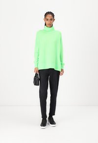 Neon grønn oversized genser med høy hals, sammen med svarte bukser og svarte sneakers, og holder en liten svart veske.
