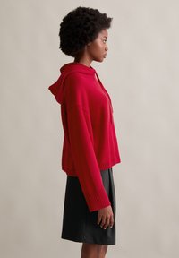 Felpa cropped rossa con coulisse e polsini a costine, abbinata a una gonna nera. Texture liscia e vestibilità rilassata valorizzano il design.