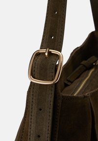 Olivově zelený semišový popruh k kabelce se zlatým čtvercovým sponou, detailním prošíváním a texturovaným povrchem. Viditelná vnitřní kapsa na zip.