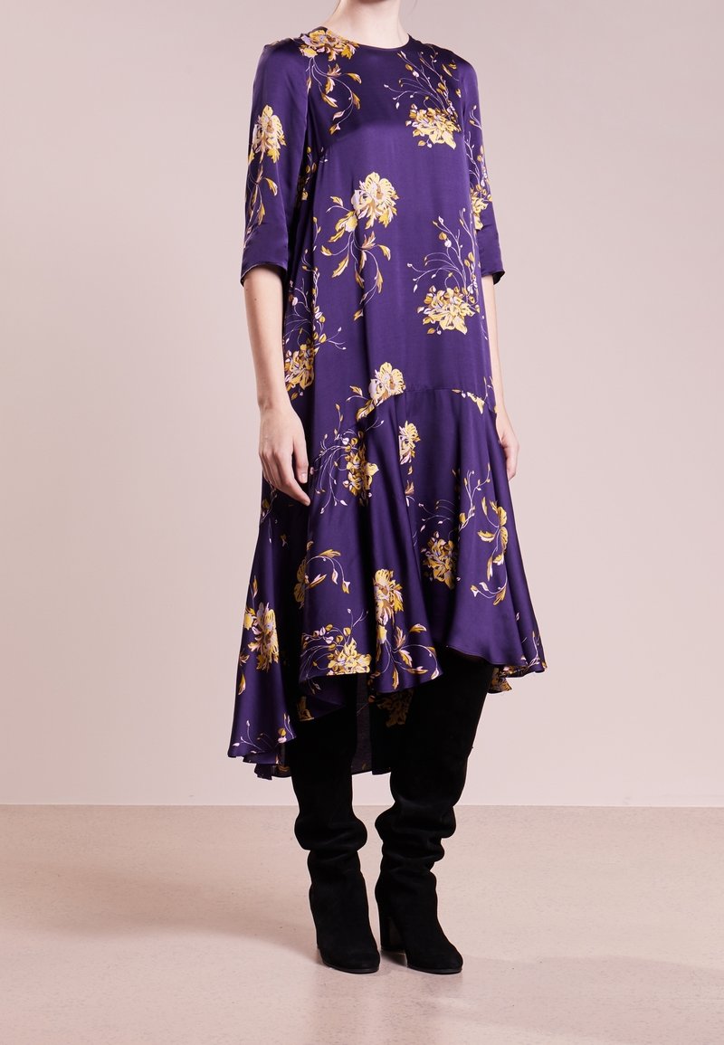 Fioletowa sukienka w kwiaty z rozkloszowanym dołem, ozdobiona żółtymi kwiatami i rękawami do łokcia, zestawiona z wysokimi czarnymi botkami.