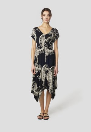 Melna kleita ar baltām smalkām rakstu detaļām, īsām piedurknēm, V-veida kakla izgriezumu, asimetrisku apakšmalu, izgatavota no mīksta auduma, kombinēta ar melnām sandālēm.