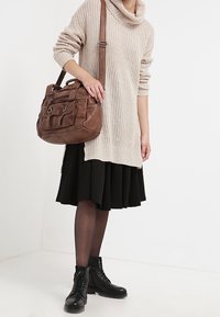 Kobieta ubrana w beżowy sweter, czarną spódnicę, przezroczyste rajstopy, czarne botki, niesie dużą brązową skórzaną torbę na ramię.