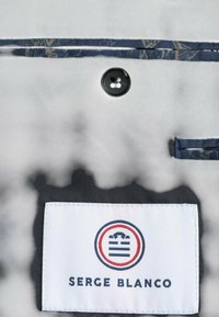 Tissu blanc avec un motif imprimé, une bande d'accent bleu foncé, un bouton et une étiquette affichant "SERGE BLANCO" avec un emblème circulaire.