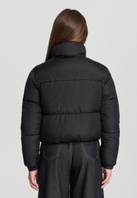 Giacca a vento nera con colletto alto, design trapuntato e polsini elastici. Indossata sopra pantaloni in denim scuro, che mettono in risalto una texture liscia.