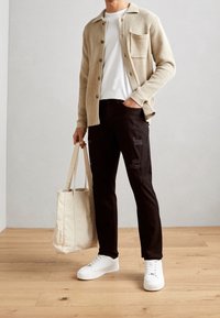 Mann in beiger Strickjacke, weißem T-Shirt, schwarzen zerrissenen Hosen und weißen Sneakers, der eine beigetönige Tasche hält und auf einem Holzboden steht.