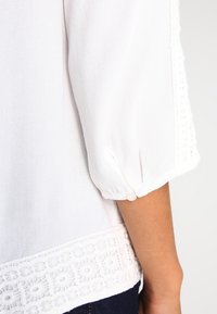Blusa bianca con maniche 3/4, orlo testurizzato con ricamo floreale e un dettaglio a bottone al polsino della manica. Tessuto morbido.