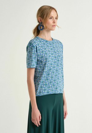 Donna con capelli biondi raccolti in una coda di cavallo, indossa una maglietta a maniche corte con motivo blu, una gonna verde scuro e grandi orecchini circolari blu, rivolta verso destra.