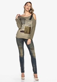 Olivegrünes schulterfreies Shirt mit gemischten Stoffeinsätzen, darunter Mesh und goldene Akzente. Kombiniert mit ausgebleichten blauen, zerrissenen Jeans.