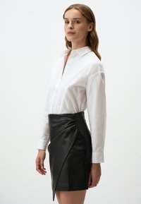 Jupe mini en cuir noir avec un design drapé et des détails froncés à la taille, associée à une chemise blanche ajustée à boutons.