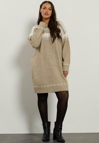 Robe en tricot beige avec un empiècement et un ourlet à motifs blancs contrastants. Présente une coupe décontractée et des poignets côtelés. Portée avec des bottines noires.