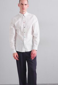 White button-up shirt with multicolored buttons, long sleeves, and a classic collar, paired with dark tailored trousers. Smooth fabric texture.