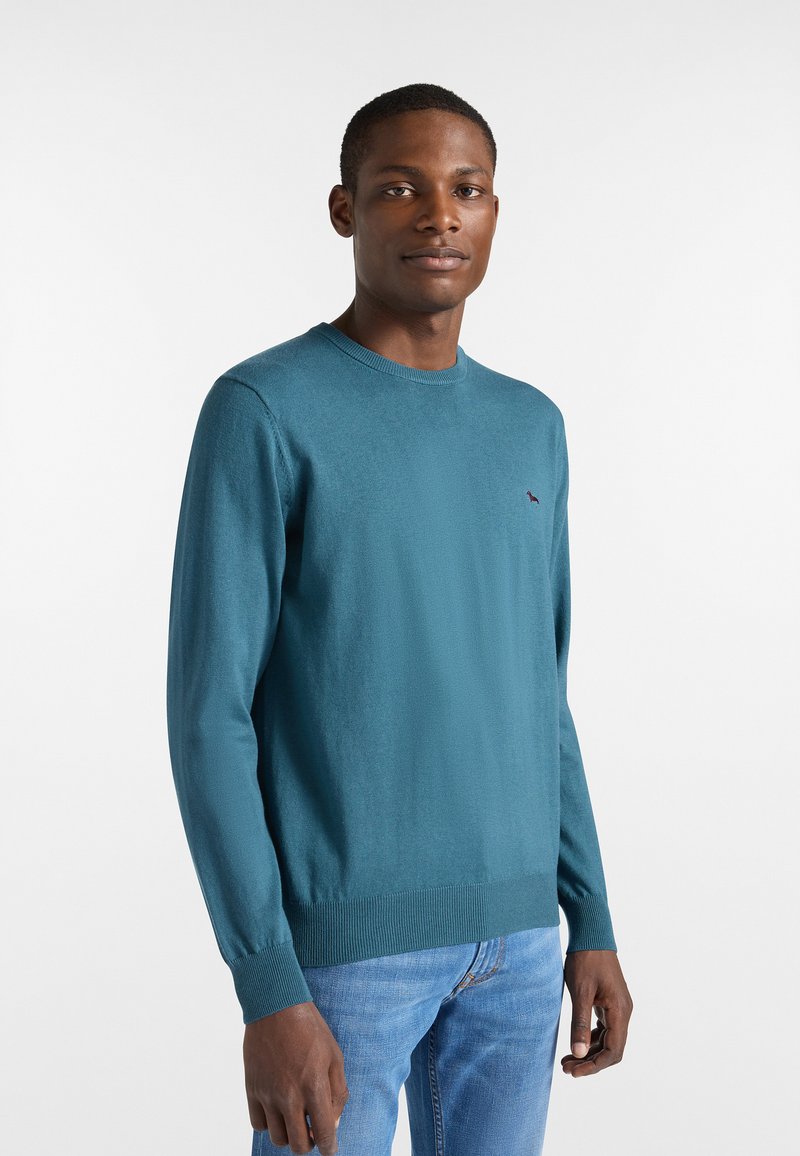 Türkisfarbener Strickpullover mit langen Ärmeln und gerippten Bündchen. Verfügt über ein kleines Logo-Detail auf der Brust. Kombiniert mit hellblauen Jeans.