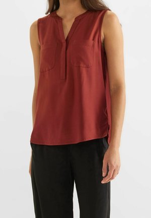 Vrouw draagt een mouwloze roestrood gekleurde blouse met twee borstzakken en een zwarte broek, staande tegen een effen witte achtergrond.