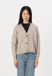 Beige gebreide cardigan met een V-hals, drie grote knopen, geribde mouwen en zoom. De textuur lijkt zacht en donzig. Gepaard met blauwe jeans.
