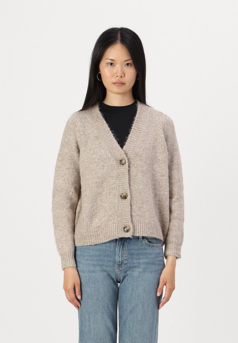 Beige gebreide cardigan met een V-hals, drie grote knopen, geribde mouwen en zoom. De textuur lijkt zacht en donzig. Gepaard met blauwe jeans.