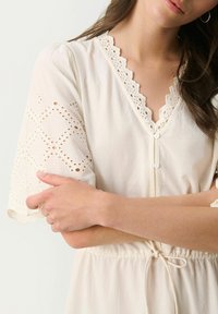 Light beige dress featuring a lace trim v-neckline, short puff sleeves with eyelet details, and a cinched waist with a drawstring.