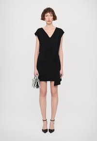 Robe noire courte avec des manches courtes, décolleté en V et un détail torsadé sur le côté, associée à des escarpins noirs pointus et un sac à main transparent.