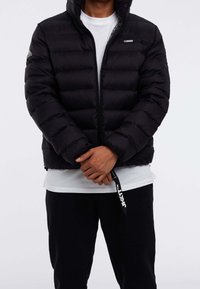 Zwarte pufferjas met horizontaal doorgestikt patroon, ritssluiting aan de voorkant en logodetail op de linkerborst. Draagt over een wit shirt en zwarte broek.