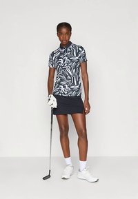 Πολό μπλούζα γκολφ με ναυτικό και λευκό αφηρημένο σχέδιο, κοντά μανίκια, γιακά, συνδυασμένη με μαύρη φούστα. Λευκό γκολφ γάντι και παπούτσια.