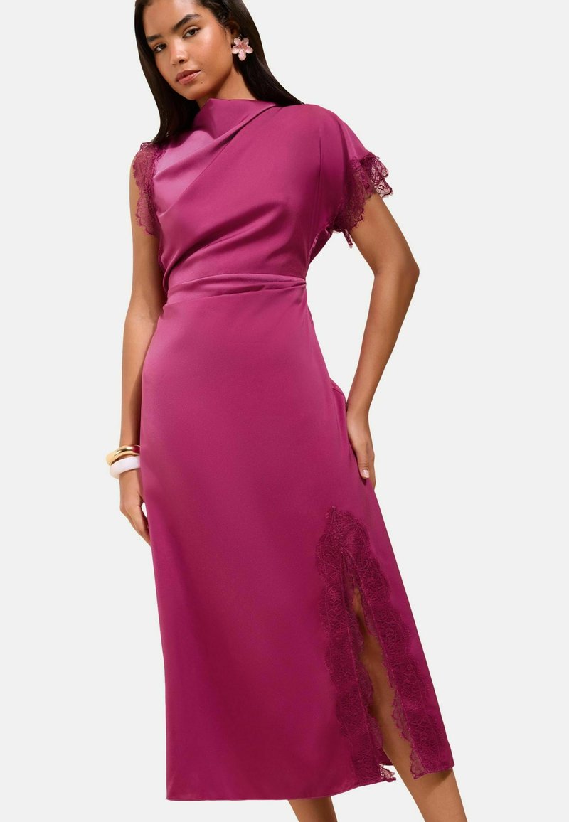 Woman wearing a sleeveless magenta dress with lace trim on one shoulder and a lace slit on the skirt, accessorized with bracelets and flower earrings.