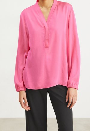 Kobieta ubrana w różową bluzkę z długim rękawem, dekoltem w serek i elastycznymi mankietami, połączoną z czarnymi spodniami, stoi na jednolitym tle.