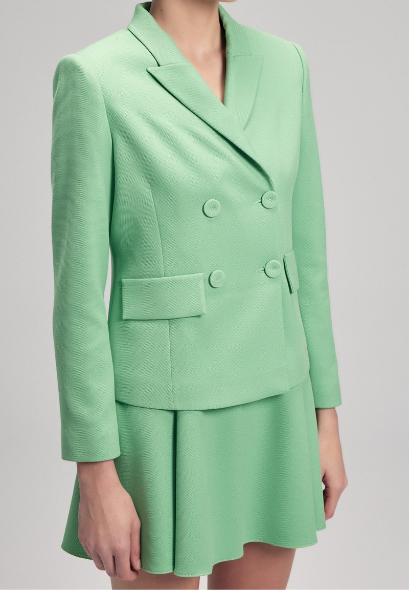 Femme portant un blazer croisé vert clair avec une jupe taille haute évasée assortie sur un fond neutre.