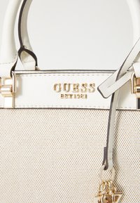 Beige og creme håndtaske med tekstureret overflade, guldfarvet hardware og prægede "GUESS EST. 1981" logo. Har korte hvide håndtag og charm.