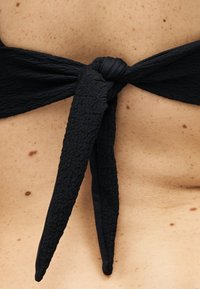 Primer plano de una tela negra texturizada atada en un lazo sobre piel humana con pecas, probablemente parte de un traje de baño o cierre de ropa.
