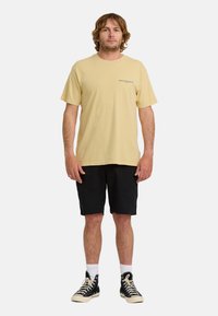 Mężczyzna z włosami do ramion, ubrany w beżowy T-shirt, czarne szorty, białe skarpetki i czarne sneakersy za kostkę, stojący na białym tle.