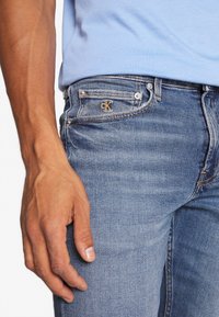 Jeans i medelblått denimmaterial med en lätt urtvättning, med en guldbroderad "ck"-logotyp på fickan och klassisk femficksdesign.