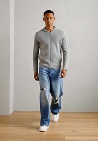 Grå hettejakke med glidelås laget av myk stoff, kombinert med lyseblå slitte jeans og hvite joggesko. Modell som står mot en nøytral bakgrunn.