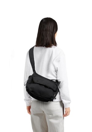 Personne portant un sweat-shirt et un pantalon gris clair avec un sac bandoulière noir sur le dos, tournée de dos à la caméra.