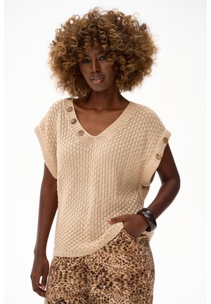 Kobieta o kręconych włosach ubrana w beżowy dzianinowy top bez rękawów z guzikami i spodnie w panterkę, pozuje z jedną ręką w kieszeni.