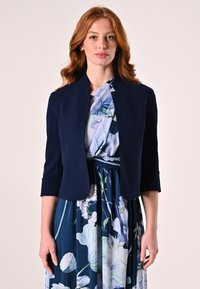Veste cropped navy à manches 3/4, texture lisse. Portée par-dessus une robe bleue à fleurs avec de grandes fleurs et des accents verts.
