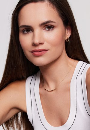 Złota naszyjnik o zakrzywionym kształcie, noszona na białej bluzce w czarne paski. Modelka ma długie, proste brązowe włosy i neutralny makijaż.
