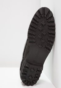 Czarna podeszwa buta z teksturowym bieżnikiem, z wzorowanym designem i wytłoczonym logo. Materiał wydaje się trwały i odpowiedni do różnych terenów.