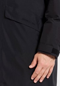 Svart vattentät jacka med texturerat tyg, kardborrejustering vid ärmslut och ett broderat varumärkeslogo på bröstfickan.