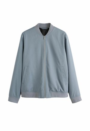 Junger Mann mit braunen Sonnenbrillen und einer hellgrauen Zip-up-Jacke, stehend im weichen natürlichen Licht mit neutralem Ausdruck.