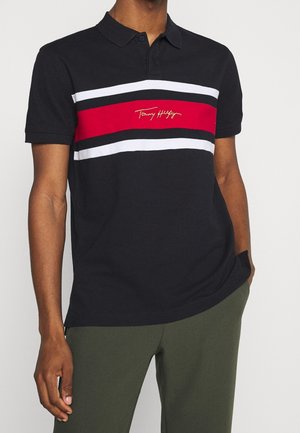 Sort polo med røde, hvide og sorte vandrette striber og et lille logo på brystet, båret af en person med grønne bukser.